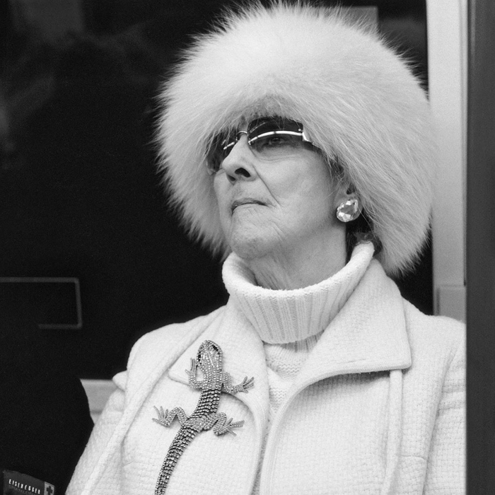 black and white photo of a older woman looking to the horizon wearing fur hat, white pullover, sun glasses and a big lizard