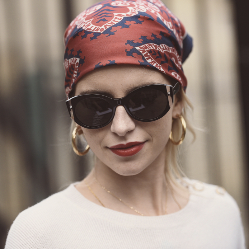 Chic woman wearing a white top, sunglasses, gold hoops and a silk scarf on her head.