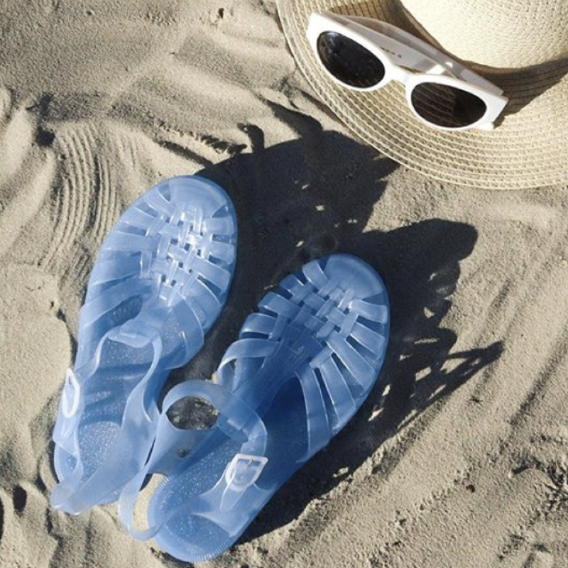 Blue jelly sandals next to sunglasses and straw hat on the sand.
