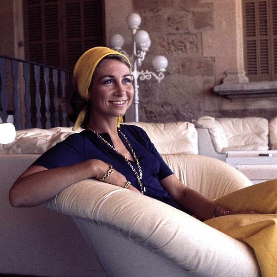 Queen Sofia of Spain sitting at a summer terrace wearing a yellow hair scarf, dark blue top and yellow trousers