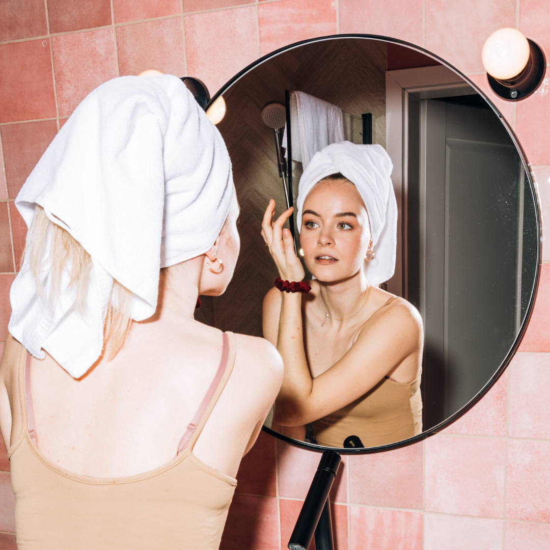 A girl applying cream to her face in her bathroom, with a toil wrapped on her hair