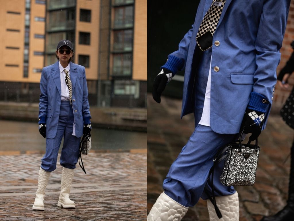Fun CPHFW 22 street style - fashionista wearing a blue pant suit, with checked tie, white boots and sparkling Prada bag