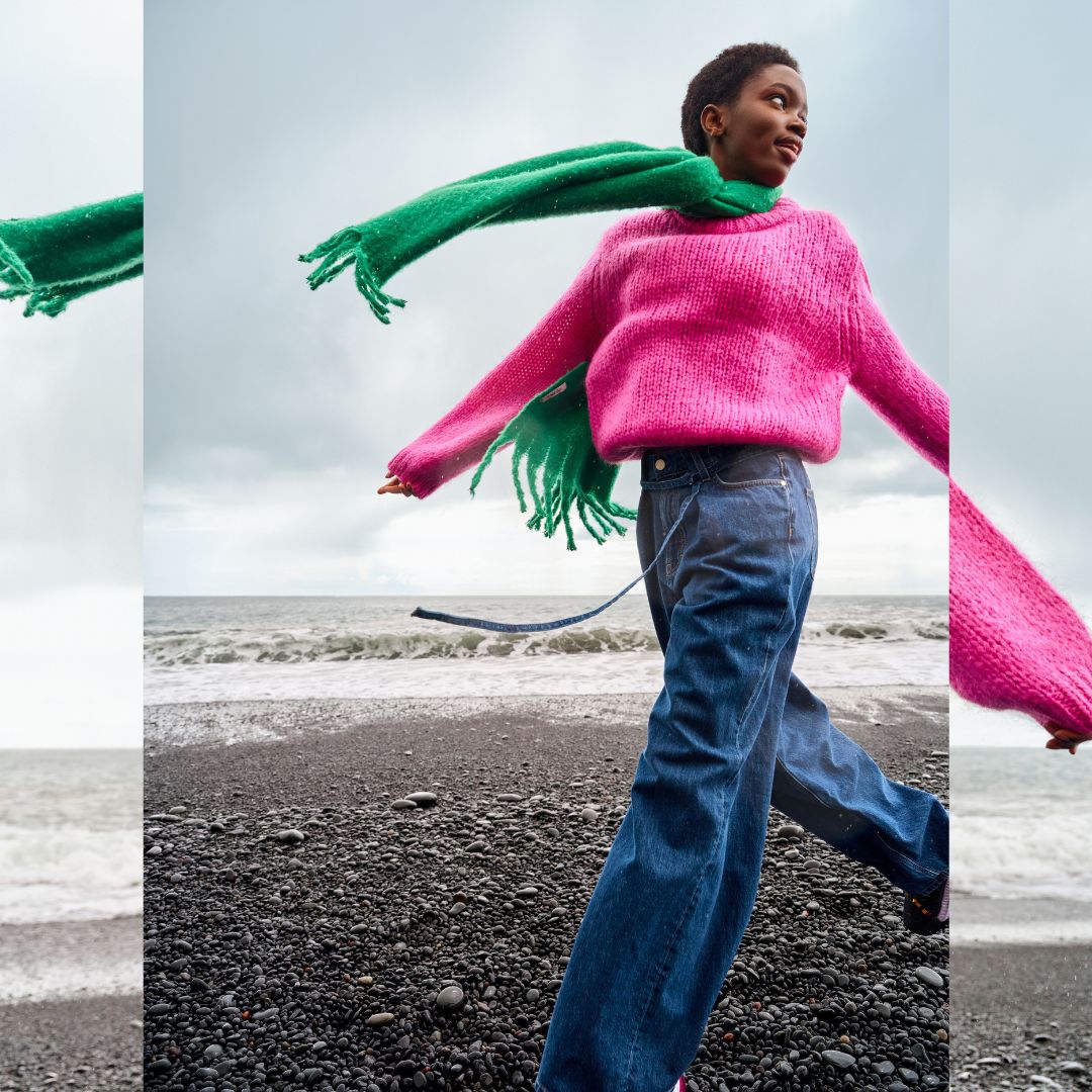 model on the sea side wearing a pink pullover with a green foulard and a denim pants