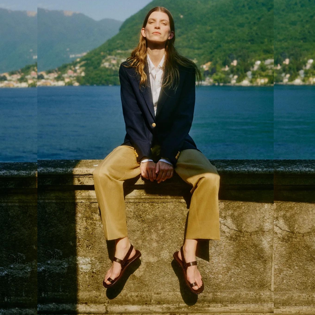 model sits on a wall on the seaside wearing pants, a blazer and a pair of sandals from Brioni.